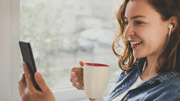 Kulaklıklı genç bir kadın video görüşmesi için akıllı telefon kullanıyor, bardağını evin içinde tutuyor. Görüntülü zoom videosu çeken kız akıllı telefondan arıyor. Sosyal medya, evde kal.