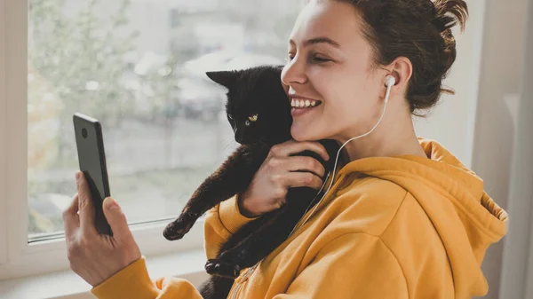 Kulaklık takan neşeli genç bir kadın elinde siyah bir kedi tutuyor. Video görüşmesi için akıllı telefon kullanıyor. Arkadaşına ya da ailesine selam veriyor. Mutlu gülümseyen hippi kız görüntülü yakınlaştırma videosu çekiyor.
