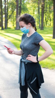 Tıbbi maske takan genç bir kadın spor sonrası sosyal medyada veri paylaşıyor, şehir parkında koştuktan sonra cep telefonundan müzik dinliyor. Karantina sırasında aktif yaşam, spor, covid-19.