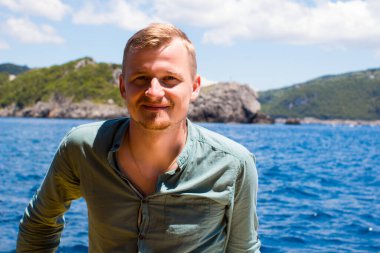 Dağ tropik koyunun arka planında mavi zümrüt kristal berrak deniz ve gökyüzü ile gülümseyen sarışın bir adamın portresi. Mutlu milenyum erkekleri sıradan tişörtler giyer.