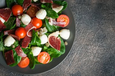 Prosciutto, mozzarella, vişneli domates, kapari ve ıspanaklı salata. Arka planda koyu bir tabakta..