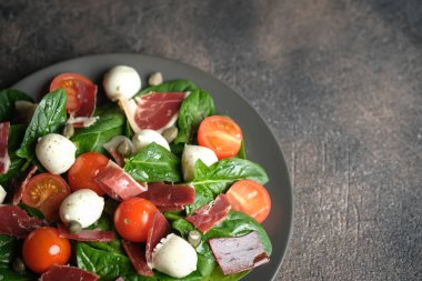 Prosciutto, mozzarella, vişneli domates, kapari ve ıspanaklı salata. Arka planda koyu bir tabakta..