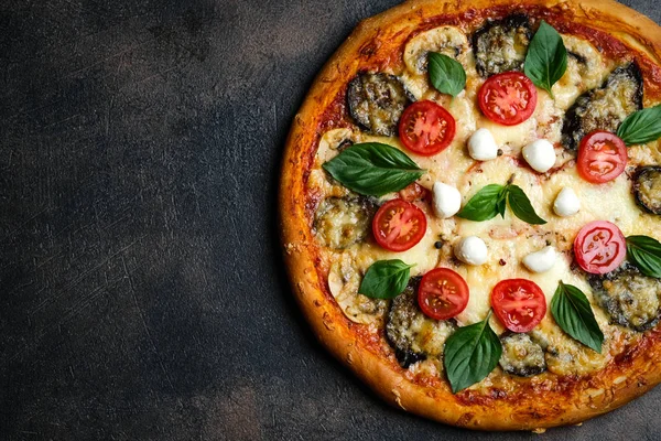 Traditional Italian pizza with eggplants, mozzarella, basil and tomatoes on a dark background top view copy space.