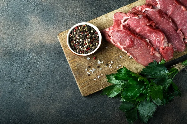 Fresh raw beef, cut into steaks  with vegetables, herbs and spices on a wooden board on a dark background. Copy space. Top view