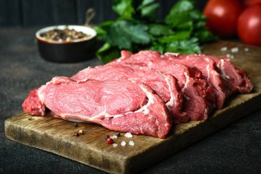 Taze çiğ et, ahşap bir tahtanın üzerinde biftekler, sebzeler, otlar ve baharatlarla karanlık bir arka planda tahta bir tahtada kesilmiş..