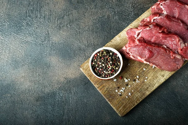 Fresh raw beef, cut into steaks on a wooden board with vegetables, herbs and spices on a wooden board on a dark background. Top view.