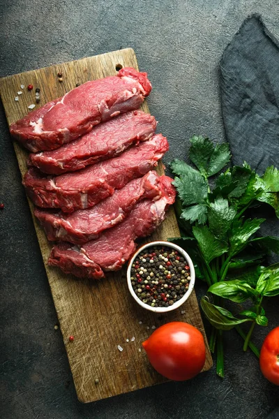 Fresh raw beef, cut into steaks on a wooden board with vegetables, herbs and spices on a wooden board on a dark background. Top view.