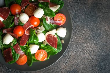 Salad with prosciutto, mozzarella, fresh vegetables on a dark plate on a black background top view copy space.