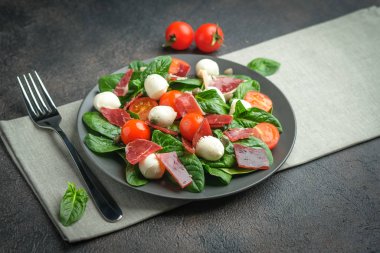 Salad with prosciutto, mozzarella, fresh vegetables on a dark plate on a black background.