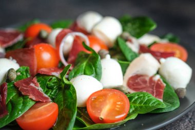 Salad with prosciutto, mozzarella, fresh vegetables on a dark plate on a black background.