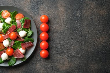 Salad with prosciutto, mozzarella, fresh vegetables on a dark plate on a black background top view copy space.