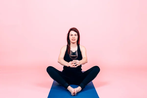 Güzel bir kadın yoga pozisyonunda oturmuş pembe arka planda bir kadeh şarap tutuyor. Şarap ve yoga konsepti.