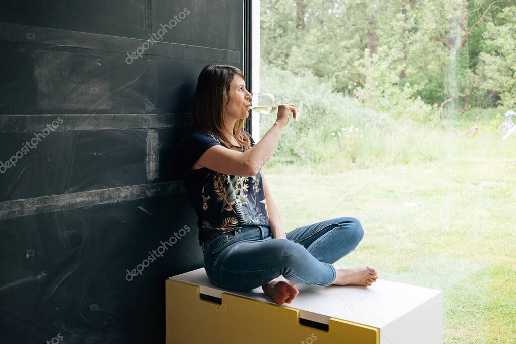 woman alone looking outside the window drinking a glass of wine on sunny day - spring summer quarantine sorrow at home