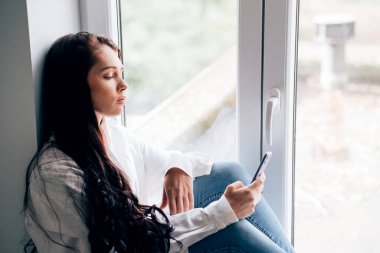 Evin penceresinde oturan genç bir kadın elinde telefon ve mesaj tutuyor.