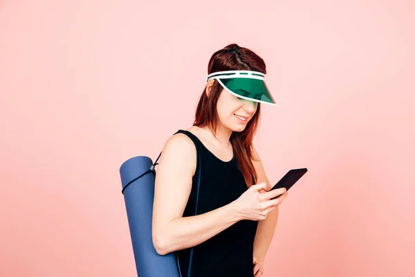 Mutlu ve gülümseyen sporcu kadın. Paspas ve güneş gözlüklü. Pembe arka planda cep telefonu kullanıyor.