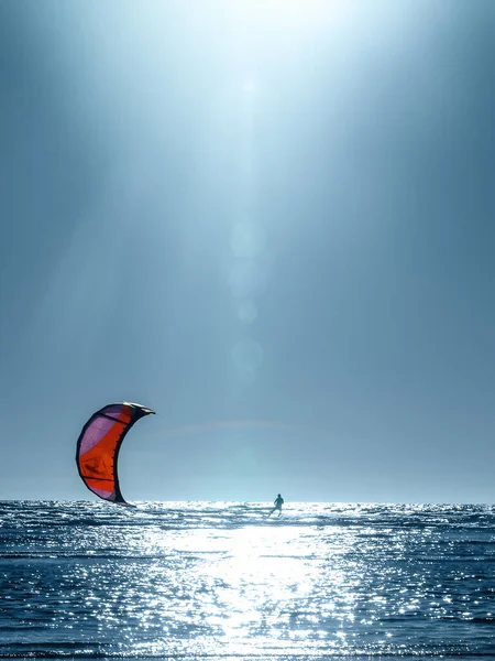 kites flies at the background of blue sea and sky, and bright sun with lens flare, and glowing light, kitesurfing  (the sail pulls along the waves of a man on the board)