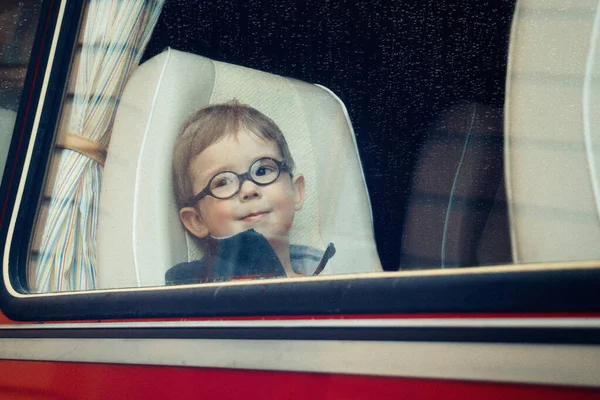 A little boy with round glasses looks through the long distance bus window with raindrops on it  (vintage processing, noises, film photo)
