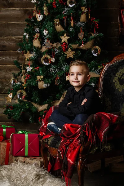 Sehr netter Junge sitzt neben einem Weihnachtsbaum und lächelt glücklich verpackt — Stockfoto