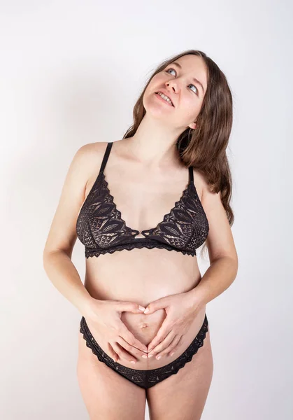 Junge schöne schwangere Frau mit langen gesunden lockigen Haaren posiert in schwarzer Unterwäsche auf grauem Hintergrund. Aktive Schwangerschaft. — Stockfoto