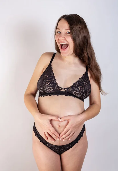 Junge schöne schwangere Frau mit langen gesunden lockigen Haaren posiert in schwarzer Unterwäsche auf grauem Hintergrund. Aktive Schwangerschaft. — Stockfoto