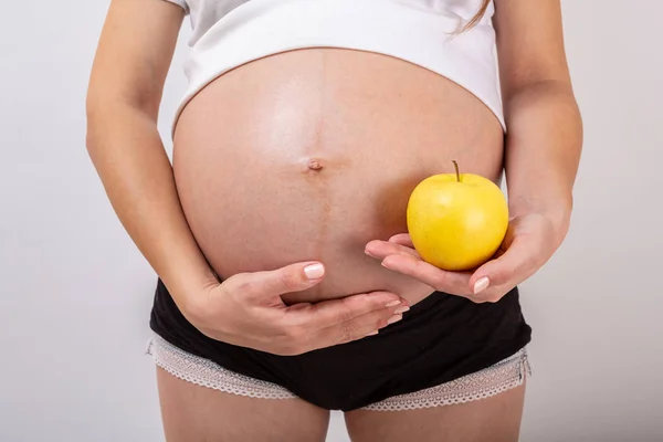 Imagen de una mujer embarazada tocando su gran vientre y sosteniendo la manzana amarilla en la mano sobre fondo blanco . — Foto de Stock