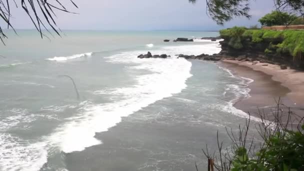 Flygburen statisk utsikt över havsytan, lugna vågor som rullar på stenig strand. Fantastiska höga klippor ovanför havet och Uluwatu templet på toppen. Bali, Indonesien — Stockvideo