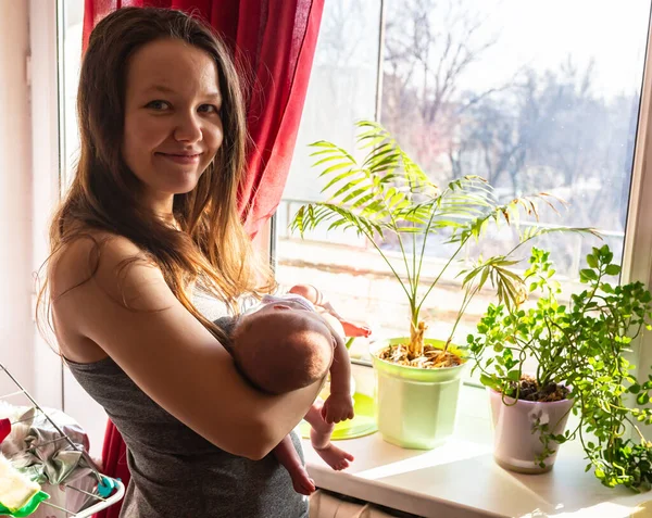 Mãe e bebê perto da janela ensolarada com flores da casa — Fotografia de Stock