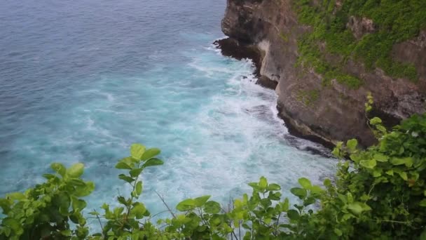 Aerial static view of evening ocean surface, calm waves rolling at rocky shore. Amazing high cliffs above the ocean and Uluwatu temple on the top. Bali, Indonesia — Stock Video
