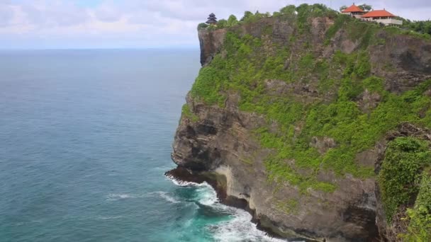 Flygburen statisk utsikt över havsytan, lugna vågor som rullar på stenig strand. Fantastiska höga klippor ovanför havet och Uluwatu templet på toppen. Bali, Indonesien — Stockvideo
