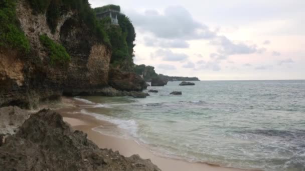 Statische Luftaufnahme der abendlichen Ozeanoberfläche, ruhige Wellen, die am felsigen Ufer rollen. Erstaunlich hohe Klippen über dem Ozean und oben der Uluwatu-Tempel. Bali, Indonesien — Stockvideo