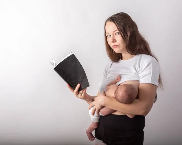 Bela jovem mãe amamentando um bebê enquanto lê um livro preto — Fotografia de Stock