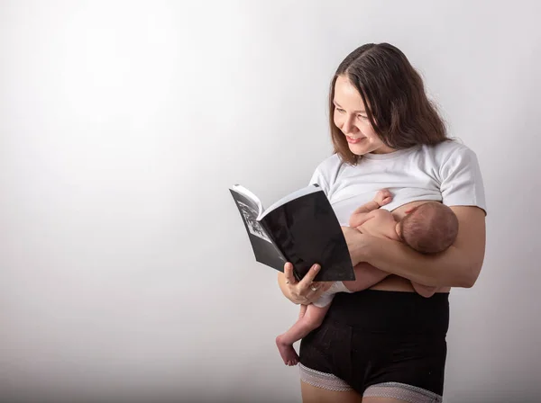 Bela jovem mãe amamentando um bebê enquanto lê um livro preto — Fotografia de Stock