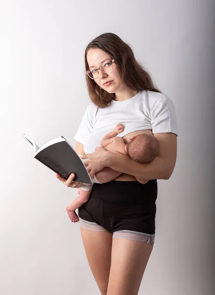 Bela jovem mãe amamentando um bebê enquanto lê um livro preto — Fotografia de Stock