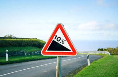 Red triangle road sign indicating a steep 10 percent downhill gradient in the road ahead. Empty road with crash barriers surrounded by green grass in the background. Traffic signs clipart