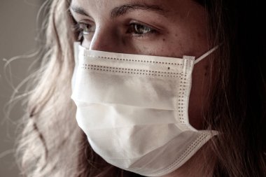 Beyaz bir tıbbi maske takan genç beyaz bir kadının ayrıntıları. Yüzün ön kısmına odaklan, arka plan bulanık. Coronavirus, COVID-19 karantinası. Koruma için maske. Doktor, hemşire konsepti.