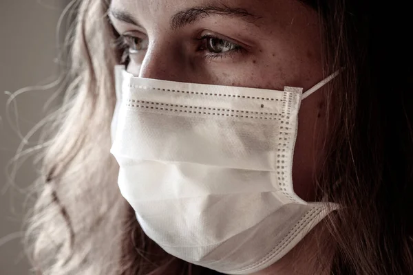 Detail of young Caucasian woman wearing a white medical face mask. Focus on the front part of the face, blurred background. Coronavirus, COVID-19 quarantine. Mask as protection. Doctor, nurse concept.