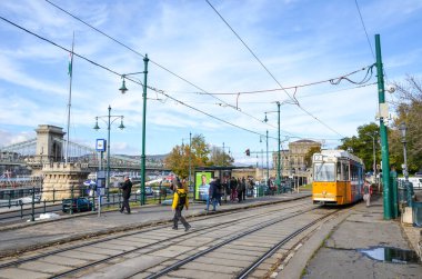 Budapeşte, Macaristan - 6 Kasım 2019: Devlet istasyonunda sarı tramvay. Etrafta dolaşan insanlar. Szechenyi zincir köprüsü, MTA binası ve arka plandaki tarihi merkez. Sokaktaki insanlar. Güneşli bir gün.