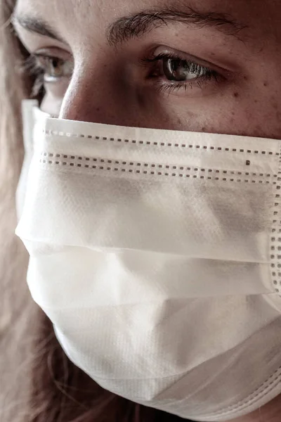 Detail of a young white woman wearing a white medical face mask. Focus on the front part of the face, blurred background. Coronavirus, COVID-19 quarantine. Mask as protection. Doctor, nurse concept.