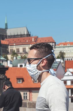 Çek Cumhuriyeti Prag 'daki Charles Köprüsü' nde güneş gözlüğü ve tıbbi maske takan genç bir adam. Arka planda bulanık eski kasaba ve Prag Kalesi. Seyahat, Coronavirus sırasında turizm. COVID-19.