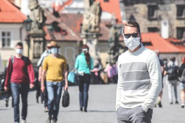 Çek Cumhuriyeti 'nin Prag kentindeki Charles Köprüsü' nde güneş gözlüklü ve dikilmiş kumaş maskeli genç bir adam fotoğraflandı. Arka planda bulanık insanlar var. Seyahat, Coronavirus sırasında turizm. COVID-19.