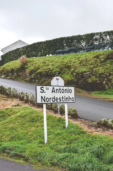 City sign Santo Antonio de Nordestinho in Sao Miguel Island, Azores, Portugal. Rural road on the hill, green grass, overcast sky. Parish, village in the Portuguese municipality. Vertical photo.