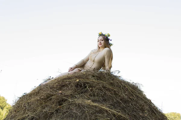 Menina loira perto de um palheiro. Uma coroa de flores na cabeça. fabricação de feno . — Fotografia de Stock