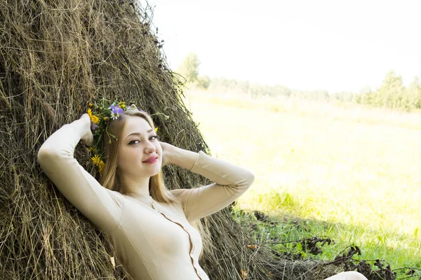 Une blonde près d'une botte de foin. Une couronne de fleurs sur la tête. fenaison . — Photo