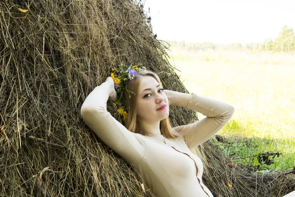 Menina loira perto de um palheiro. Uma coroa de flores na cabeça. fabricação de feno . — Fotografia de Stock