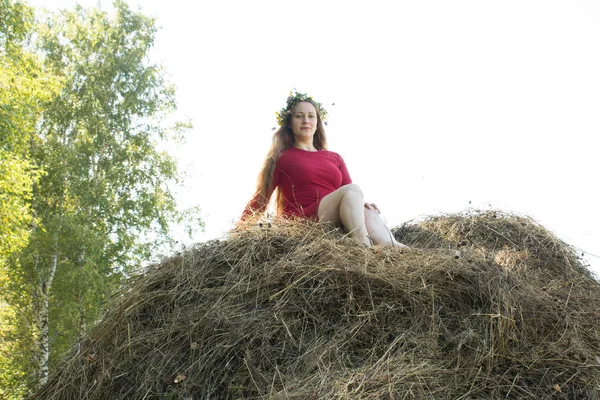 Blondýnka u kupky sena. Věnec s květinami na hlavě. senoseč. — Stock fotografie