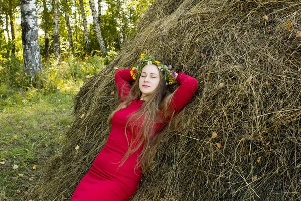 Menina loira perto de um palheiro. Uma coroa de flores na cabeça. fabricação de feno . — Fotografia de Stock