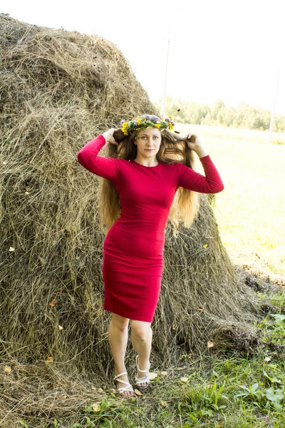 Menina loira perto de um palheiro. Uma coroa de flores na cabeça. fabricação de feno . — Fotografia de Stock