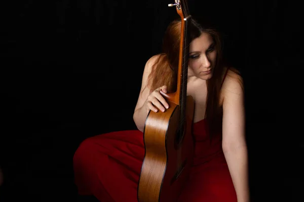 Chica en un vestido rojo se sienta en el suelo cerca de la guitarra sobre un fondo negro — Foto de Stock