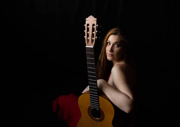Menina em um vestido vermelho senta-se no chão perto da guitarra em um fundo preto — Fotografia de Stock