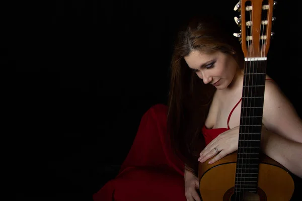 Menina em um vestido vermelho senta-se no chão perto da guitarra em um fundo preto — Fotografia de Stock